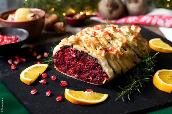 KI generiert: Das Bild zeigt eine vegetarische eingepackte Rote-Bete-Pastete, die mit Rosmarin und Granatapfelkernen garniert ist. Auf einem schwarzen Tablett sind ebenfalls Orangenstücke verteilt.