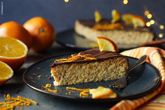 KI generiert: Das Bild zeigt ein Stück Kuchen auf einem Teller, dekoriert mit einer Scheibe Orange und Schokoladenglasur. Im Hintergrund sind weitere Kuchenstücke und ganze Orangen zu sehen.