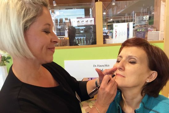 KI generiert: Auf dem Bild ist eine Kosmetikerin zu sehen, die einer Person Make-Up aufträgt. Im Hintergrund befinden sich kosmetische Produkte und Schilder.