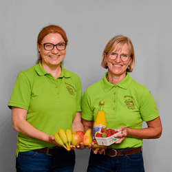 KI generiert: Das Bild zeigt zwei lächelnde Frauen in grünen Polohemden, die Obst und eine Flasche Saft in den Händen halten. Sie präsentieren frische Lebensmittel und scheinen Teil eines lokalen Marktes oder Obststands zu sein.