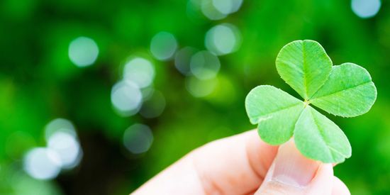 KI generiert: Das Bild zeigt eine Hand, die ein dreiblättriges Kleeblatt hält, vor einem verschwommenen Hintergrund aus üppigem Grün. Der Fokus liegt auf dem Kleeblatt, das zentral positioniert ist.