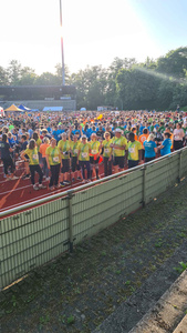 KI generiert: Das Bild zeigt eine große Gruppe von Menschen, die auf einer Laufstrecke versammelt sind, wahrscheinlich für ein Laufsportereignis. Die Teilnehmer tragen bunte Sportkleidung und sind vor einer Tribüne zu sehen.