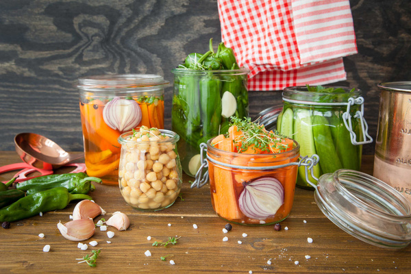 KI generiert: Das Bild zeigt eine Sammlung von Einmachgläsern, die mit verschiedenen eingelegten Gemüsesorten wie Karotten, Gurken und Paprika gefüllt sind. Im Hintergrund sind weitere Zutaten und Küchenutensilien zu sehen.