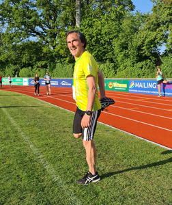 KI generiert: Das Bild zeigt eine Person in Sportkleidung, die sich auf einem Laufplatz dehnt. Im Hintergrund sind andere Läufer und eine grüne Umgebung mit Bäumen sichtbar.