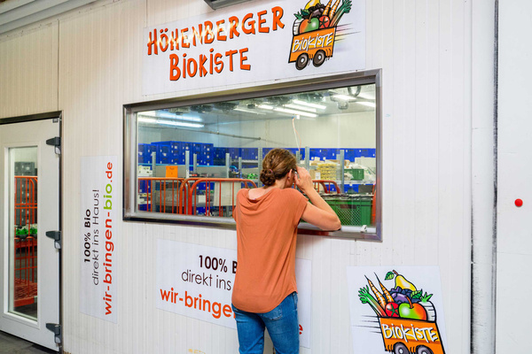 KI generiert: Ein Mann schaut durch ein Fenster in einen Raum mit Lebensmitteln bei der "Höhenberger Biokiste". Auf dem Schild steht, dass es sich um einen Lieferdienst für Bio-Produkte handelt.
