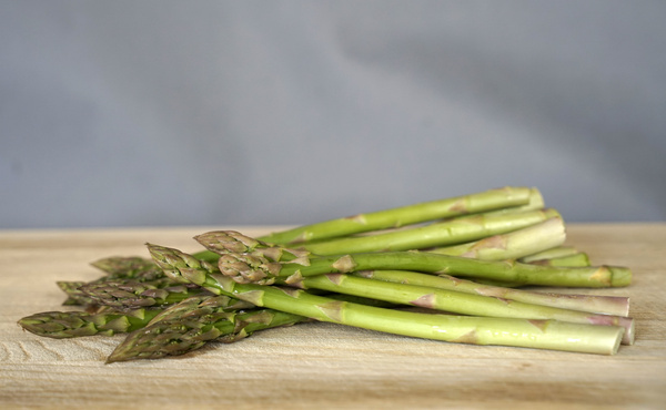 KI generiert: Ein Bündel grüner Spargelstangen auf einem Holzbrett, grauer Hintergrund.