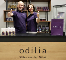 KI generiert: Zwei Personen in lila Hemden lächeln und zeigen Daumen hoch. Text: "odilia - Süßes aus der Natur".