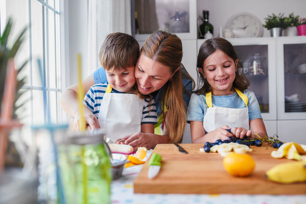 KI generiert: Eine Frau und zwei Kinder kochen zusammen in einer Küche. Sie tragen Schürzen und bereiten Obst auf einem Schneidebrett vor.