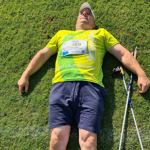 KI generiert: Ein Mann liegt erschöpft auf einer Wiese mit geschlossenen Augen, neben ihm liegen Nordic Walking-Stöcke. Er trägt ein leuchtend gelbes T-Shirt und hat eine Startnummer an der Brust.