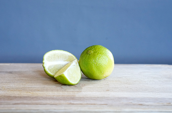 KI generiert: Eine ganze Limette und zwei Limettenscheiben auf einem Holztisch vor blauem Hintergrund.