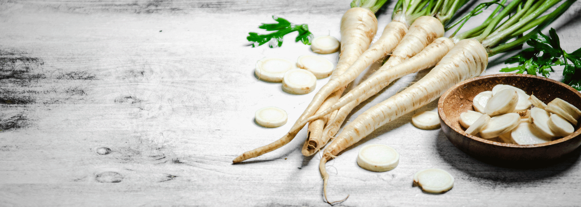KI generiert: Das Bild zeigt mehrere Pastinaken, frische und in Scheiben geschnittene, auf einer hellen Holzoberfläche. Daneben liegen einige grüne Blätter zur Dekoration.