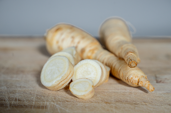 KI generiert: Das Bild zeigt geschnittene und ganze Wurzeln der Pastinake auf einem Stück Stoff, mit frischem Grün im Hintergrund. Der Hauptinhalt sind die Pastinaken samt ihrem Laub.