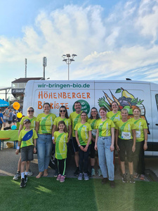 KI generiert: Eine Gruppe von Menschen in grünen T-Shirts steht vor einem Lieferwagen, der für die "Höhenberger Biokiste" wirbt. Sie scheinen bei einem Event oder Lauf teilzunehmen.