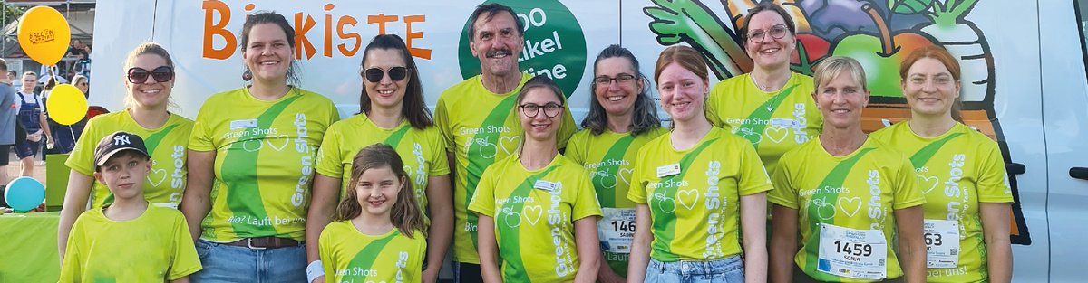 KI generiert: Eine Gruppe von Menschen in grünen T-Shirts steht vor einem bunt bemalten Hintergrund mit Gemüse und dem Schriftzug "Biokiste". Sie lächeln und tragen Starnummern, was auf eine Art von Veranstaltung oder Lauf hinweist.