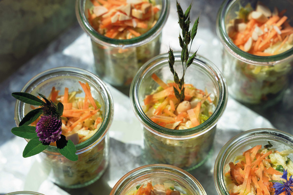 KI generiert: Auf dem Bild sind mehrere Glasgefäße mit einem bunten Salat aus Karottenstreifen, Kohlgemüse und Nüssen zu sehen, dekoriert mit Blumen und Kräutern. Die Anordnung erzeugt ein frisches und einladendes Erscheinungsbild.