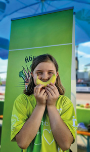 KI generiert: Ein Kind hält ein gelbes Objekt, das wie ein Lächeln geformt ist, vor seinem Gesicht. Im Hintergrund steht ein grünes Banner mit einer Illustration.